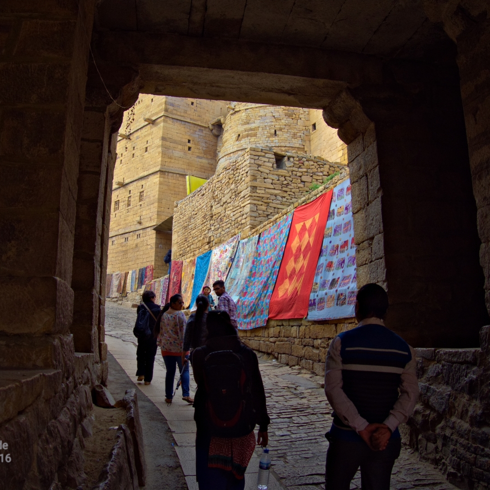 A few people are silhouetted in the arch over the narrow cobblestone street heading into the stone fort. Through the arch you can see lots of quilts and colorful textiles hanging on the stone walls.