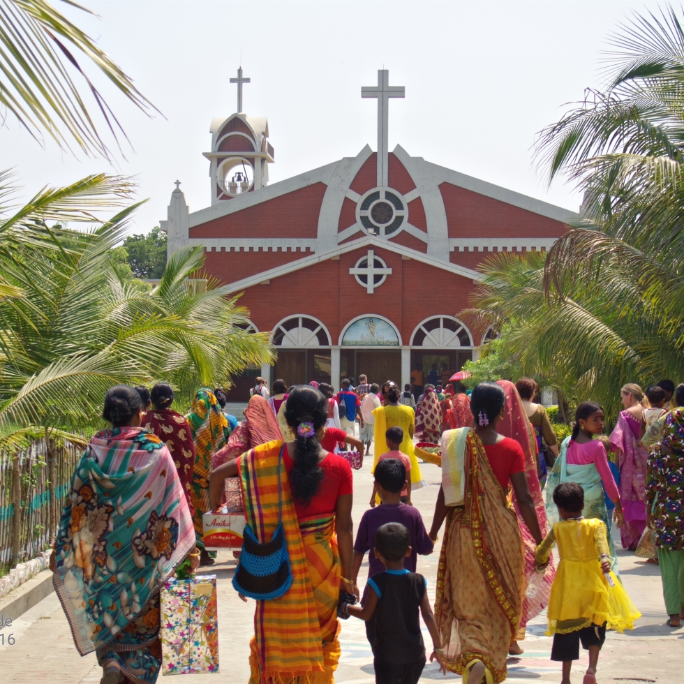 A crowd of Bangladeshi people walk away from the camera towards a church. Every one is dressed in fine clothes, especially the women who are wrapped in colorful saris. Hot sunlight shines on the big palm trees and other tropical plants lining the sidewalk.